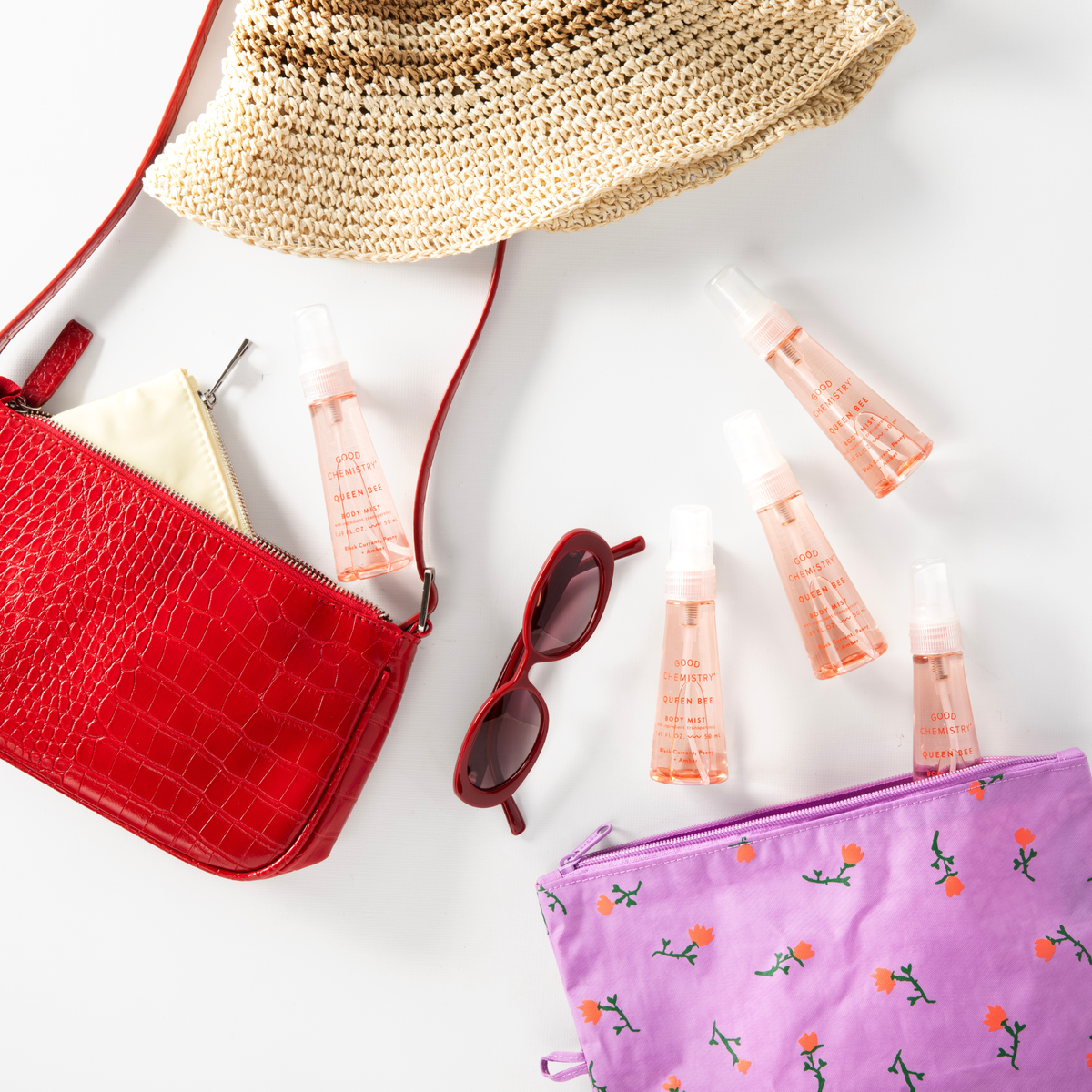 Red handbag, straw hat, sunglasses, and Queen Bee mini body mist pink bottles on a white surface