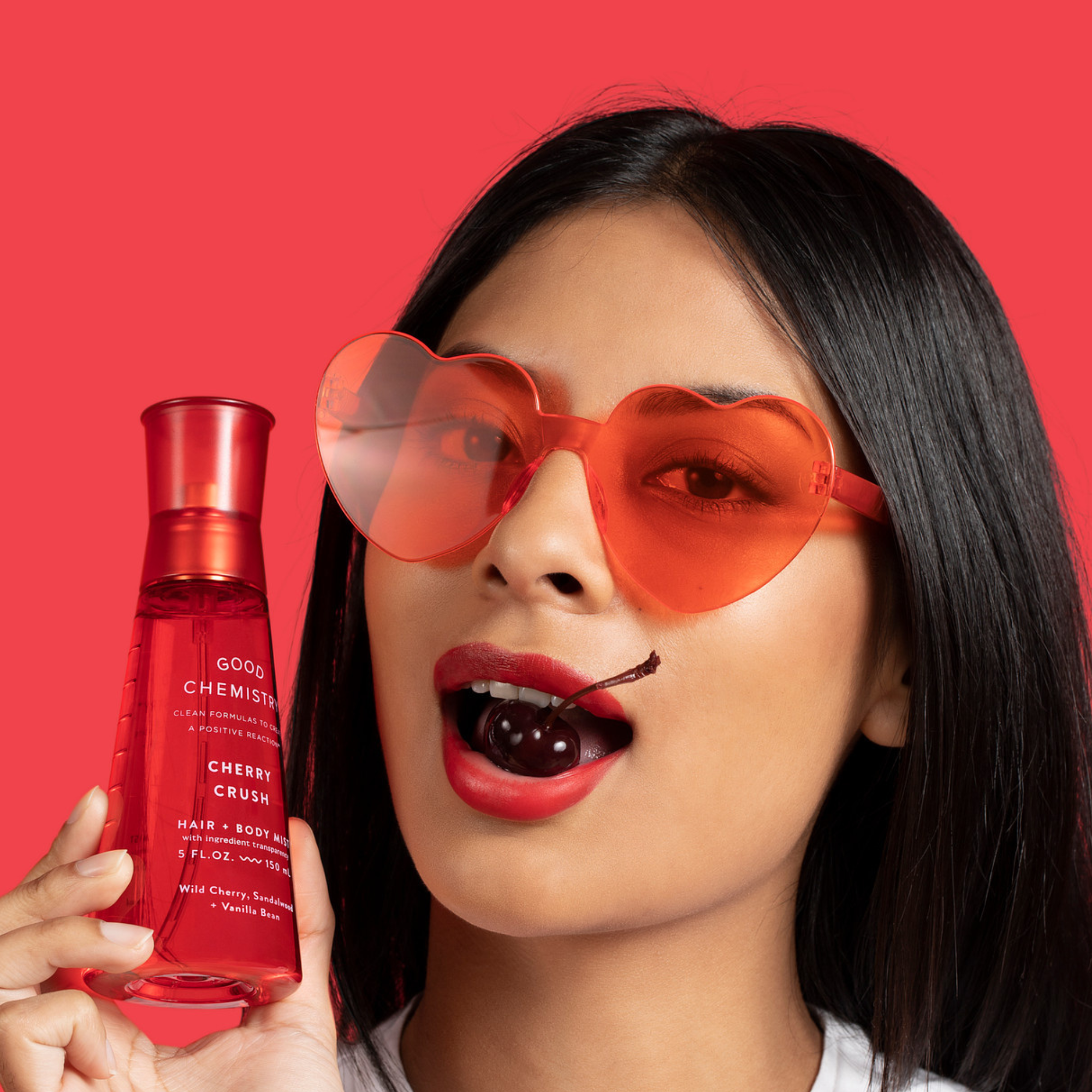Woman wearing red heart-shaped sunglasses holding 'Good Chemistry Cherry Crush'  Body Mist against a red background