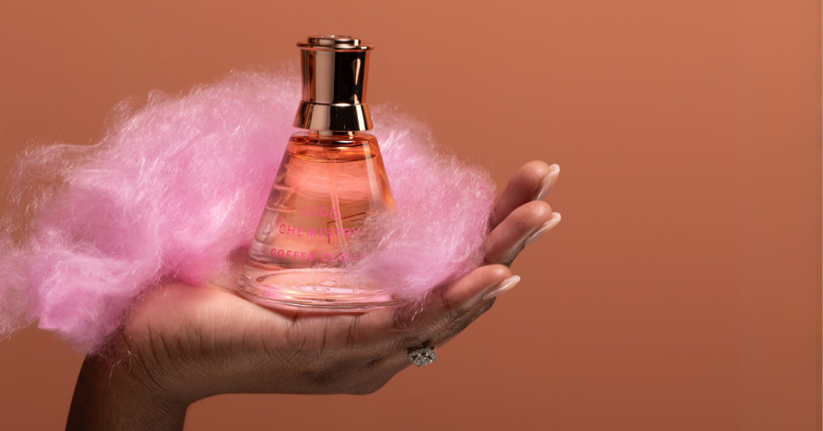 A hand holding a small perfume bottle filled with pink liquid, surrounded by fluffy pink cotton candy against a solid warm brown background.