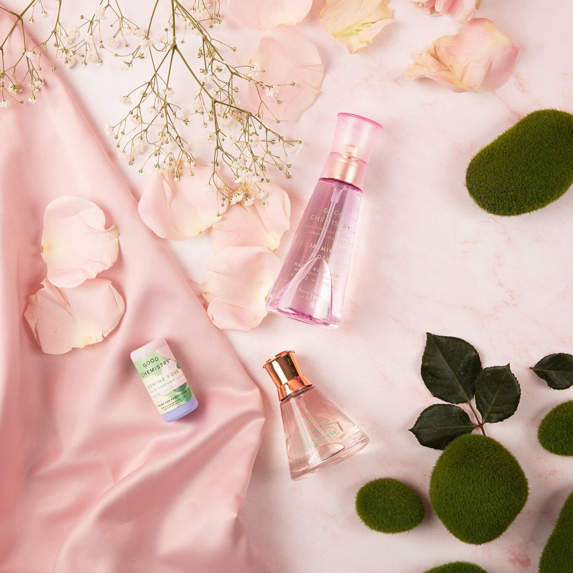 Three Jasmine Rose perfume bottles on a pink fabric background with flowers and leaves.