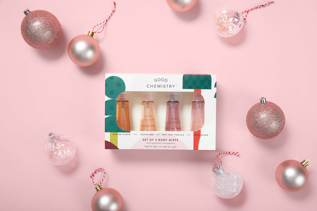 A boxed set of four mini body miststfrom Good Chemistry, displayed on a pink background with decorative pink and silver Christmas ornaments around it. The box is white with green and red graphic accents, and the lip products inside appear in peach, pink, and nude tones.