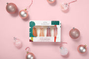 A boxed set of four mini body miststfrom Good Chemistry, displayed on a pink background with decorative pink and silver Christmas ornaments around it. The box is white with green and red graphic accents, and the lip products inside appear in peach, pink, and nude tones.