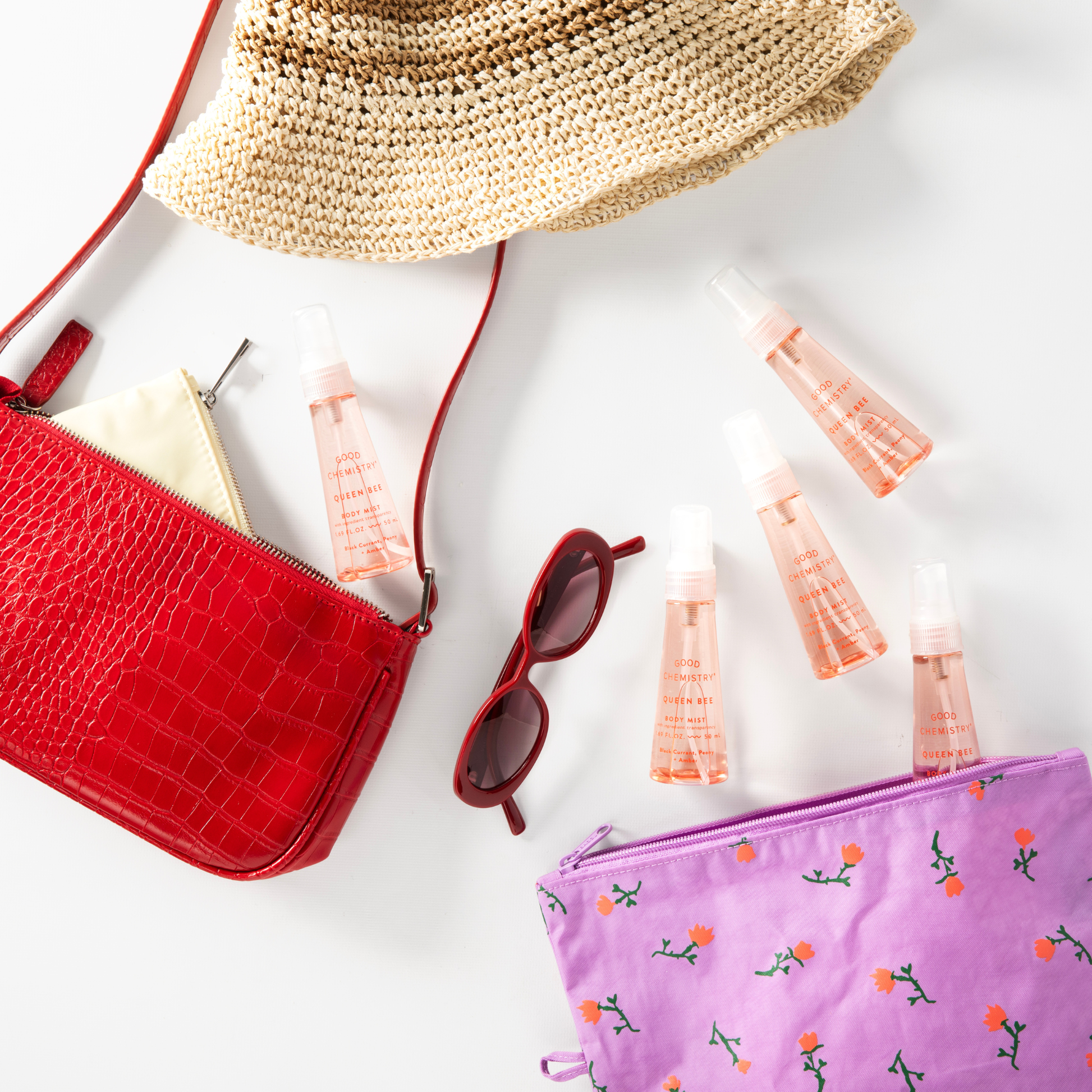 Red handbag, straw hat, sunglasses, and Queen Bee mini body mist pink bottles on a white surface
