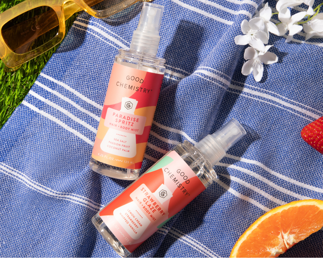 Two bottles of Good Chemistry perfume on a blue striped towel with strawberries, sunglasses, and an orange.