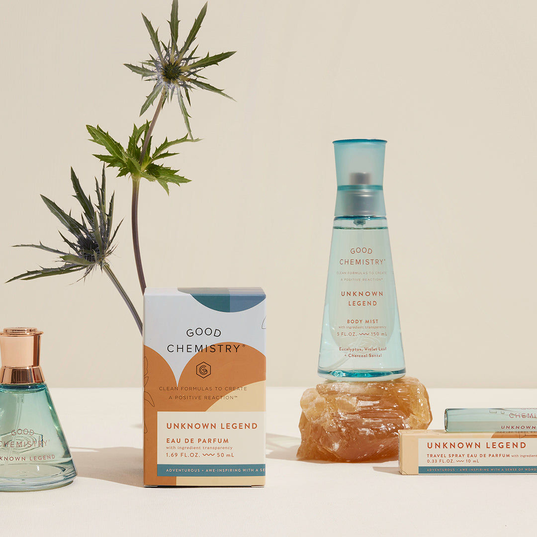 A fragrance bundle featuring a bottle of 'Unknown Legend' eau de parfum, a smaller spray bottle, and a chunk of amber-colored resin, displayed on a light surface with a plant in the background.